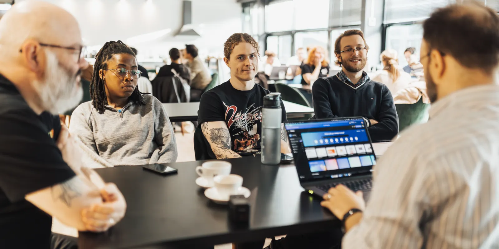 Coworkers sitting around a table with an open laptop, talking and smiling during a work meeting.