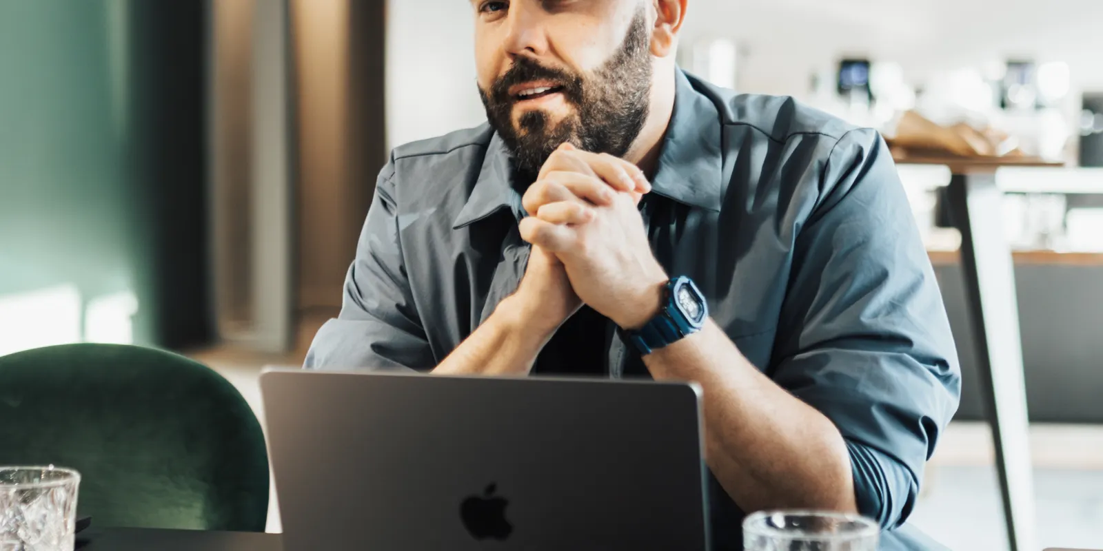 A bearded man, hands clasped in front of his laptop, listens closely during a work conversation.