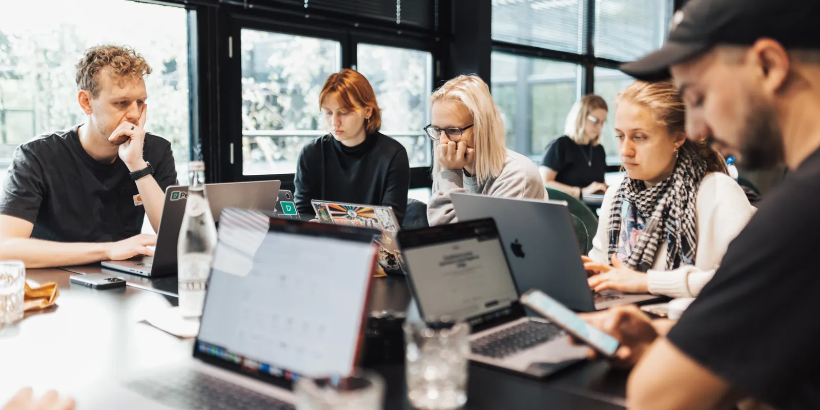 People from different teams work side by side on laptops at a long table, in front of large windows.