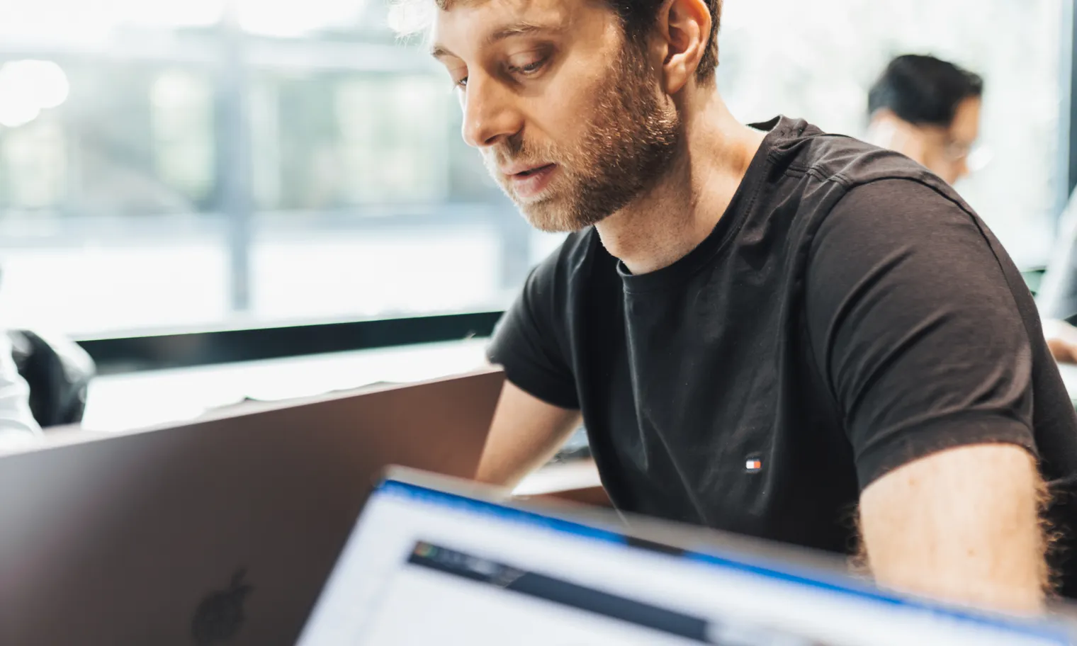 A colleague, focused, works on a laptop at a shared table, with other people in the background.