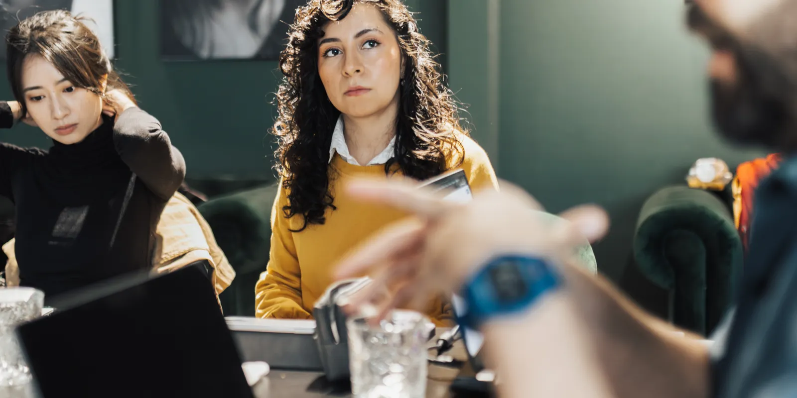 Três pessoas sentadas à mesa de uma sala de reunião ouvem alguém que gesticula ao explicar algo.