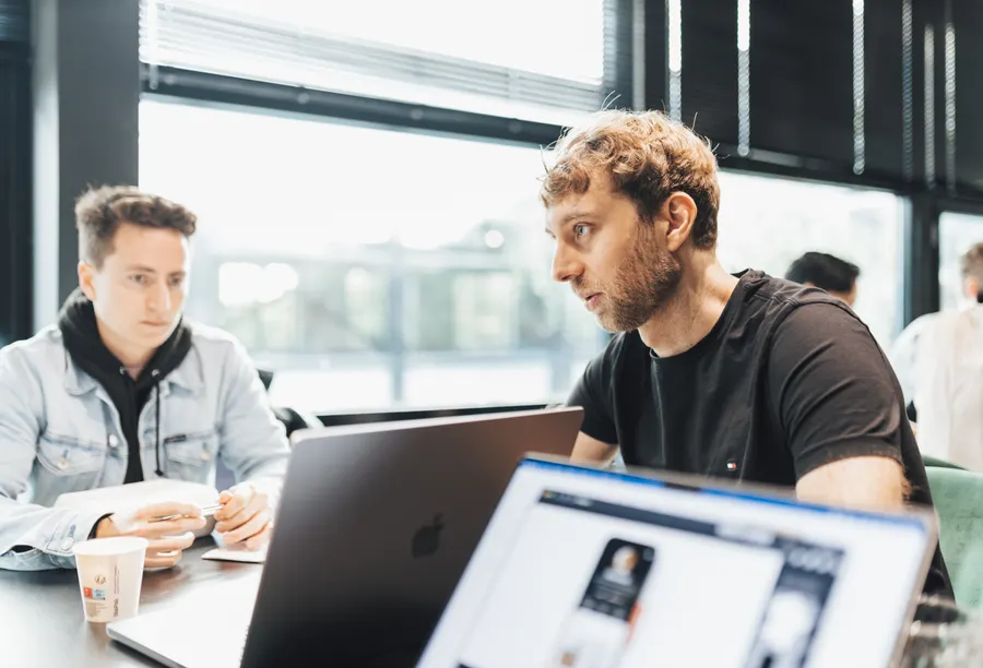Two colleagues talk in front of an open laptop at a table beside large windows.